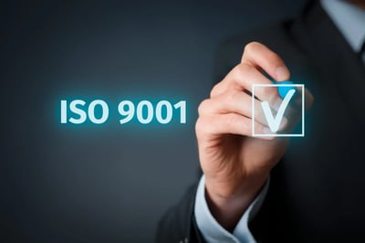Mano de persona en traje escribiendo un símbolo de verificación junto al texto ISO 9001, representando certificación de calidad.