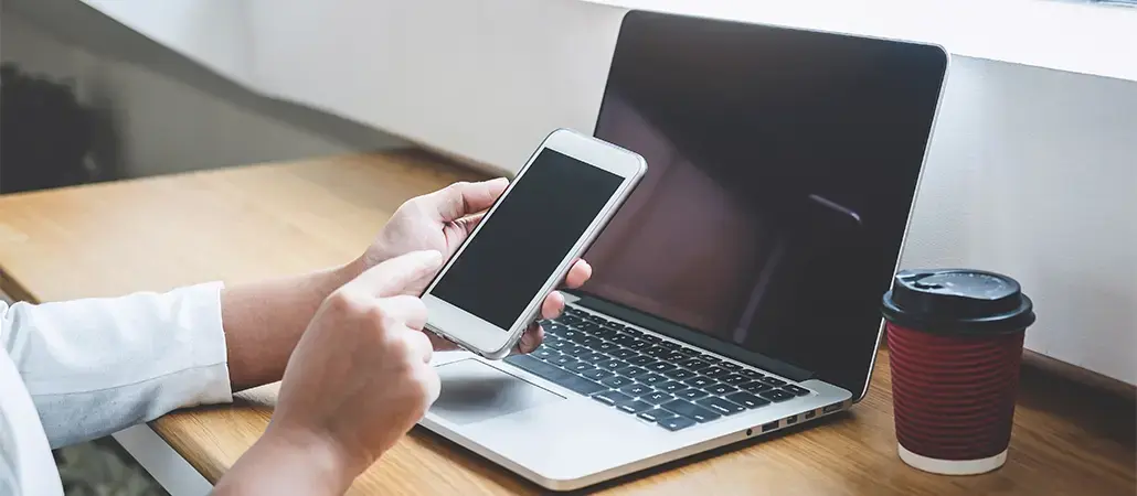 Persona usando un teléfono móvil frente a una computadora portátil en un escritorio con vaso de café.