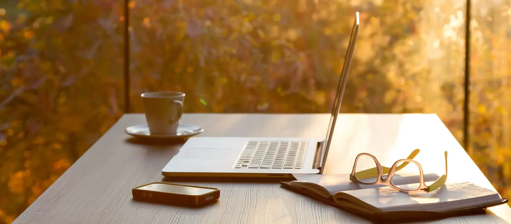 Taza de café, laptop, gafas y libreta sobre escritorio junto a ventana con luz cálida.