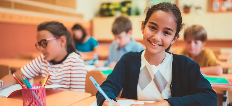 Niña sonriendo mientras escribe en su cuaderno en un salón de clases junto a otros compañeros.