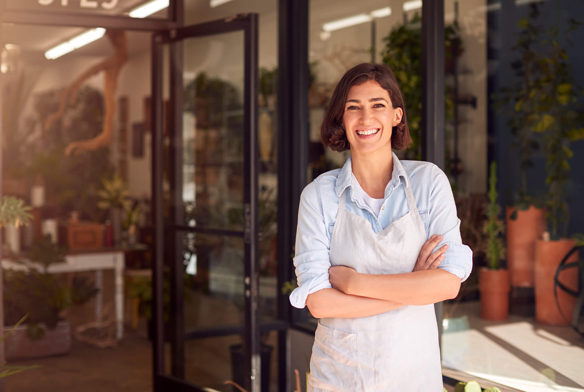 Mujer sonriente con camisa y delantal blanco de pie en la entrada de una floristería.