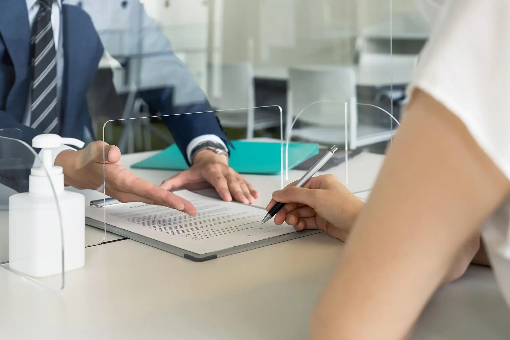 Cliente firmando un contrato en una oficina bancaria frente a un asesor financiero detrás de un acrílico protector
