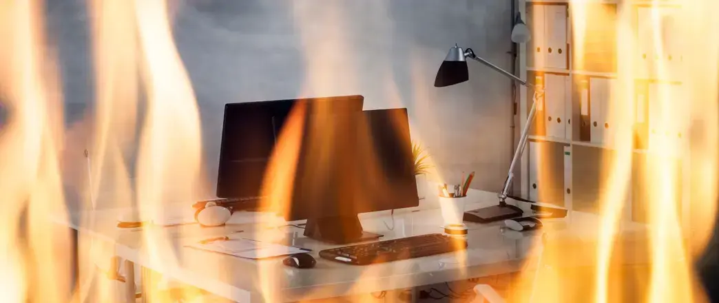 Oficina en llamas con escritorio, computadora y documentos expuestos al fuego.