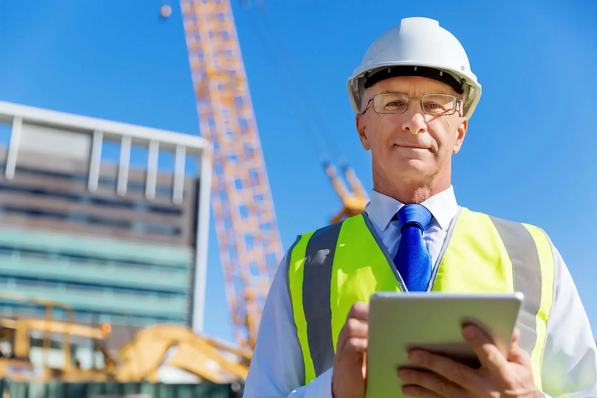 Ingeniero con casco y chaleco reflectante sosteniendo una tableta, en un sitio de construcción con grúa y edificio al fondo.