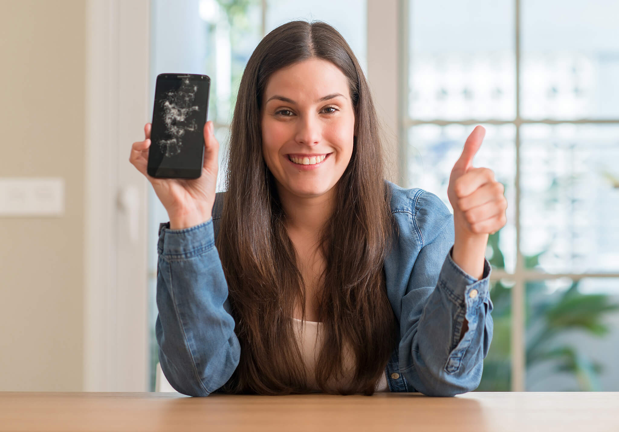 Mujer joven sonriendo, mostrando un teléfono con pantalla rota y levantando el pulgar.